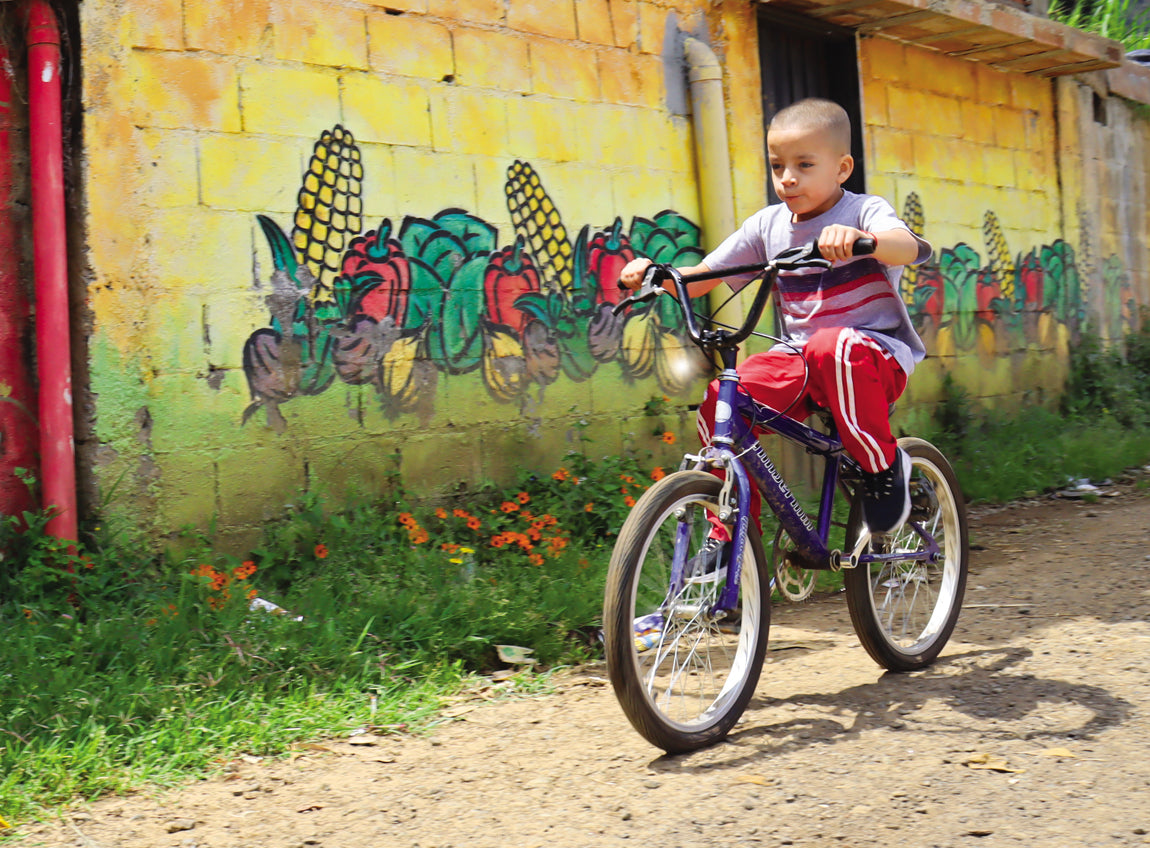  一個小男孩在巷子裡騎自行車，後面是一幅畫有蔬菜的壁畫。