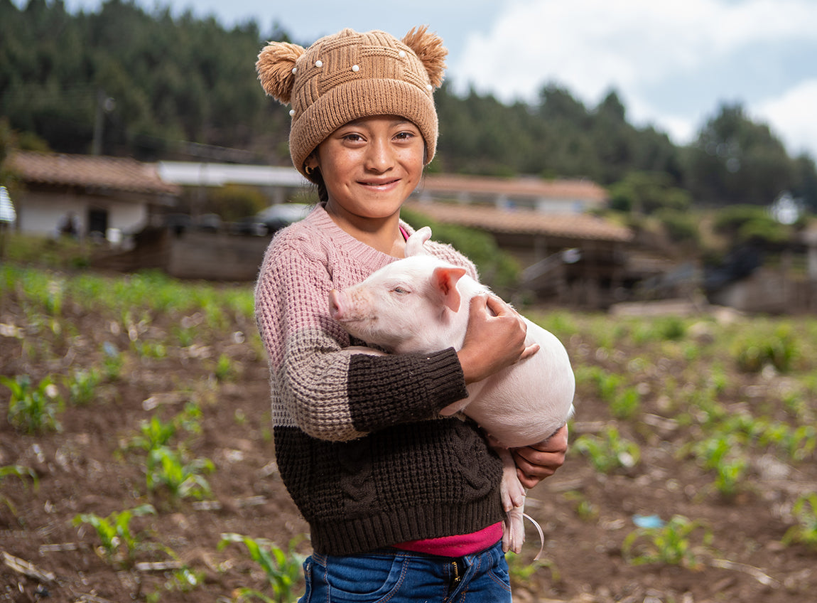  一名女童站在農地上微笑，手裡抱著一隻正在睡覺的小豬。