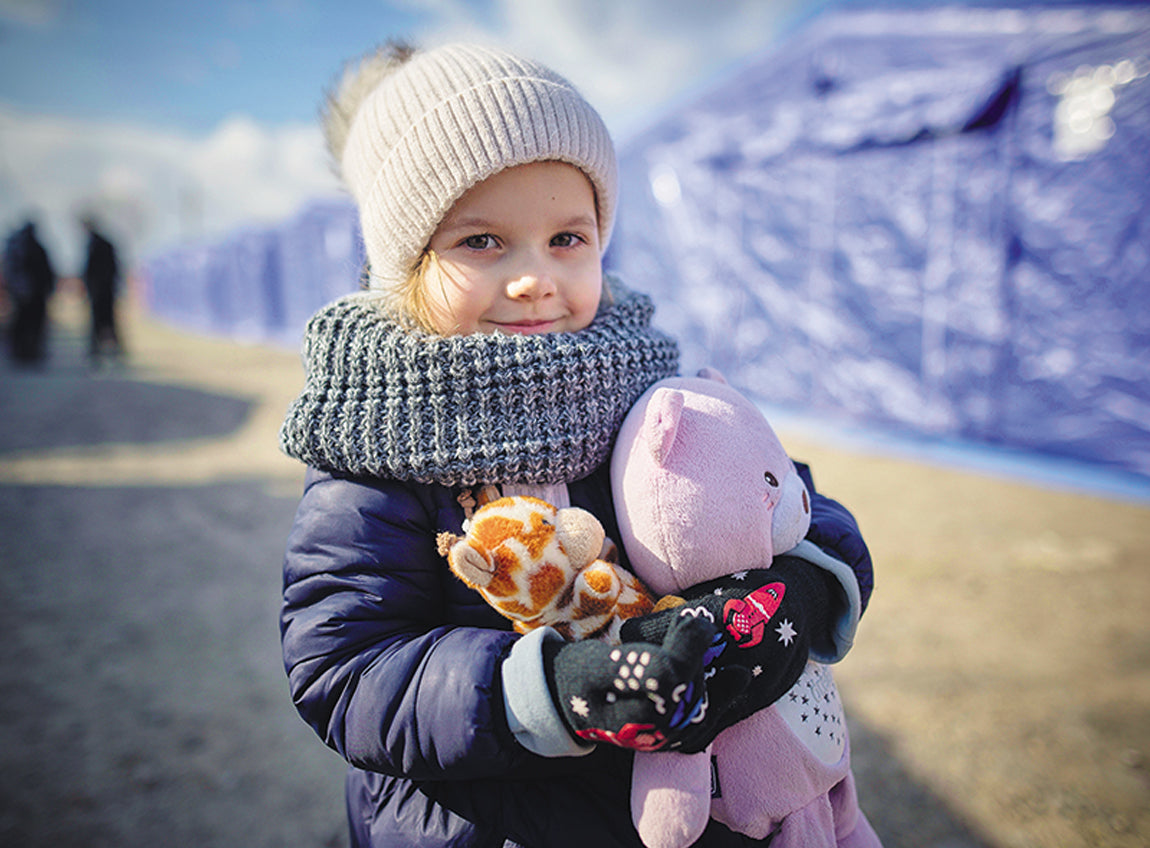  一名女童站在難民住的帳幕前，身上穿著暖和的衣服，手裡摟著一些毛絨動物。