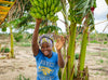 一個年青女孩微笑著向鏡頭揮手，同時抓住樹上的一束香蕉。