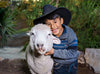 一個頭戴黑色牛仔帽的小男孩，微笑著擁抱一隻綿羊。