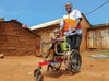一名男士身穿橙白色的世界宣明會背的，微笑著在泥路上推一張輪椅，其上坐著一個微笑著的孩子。