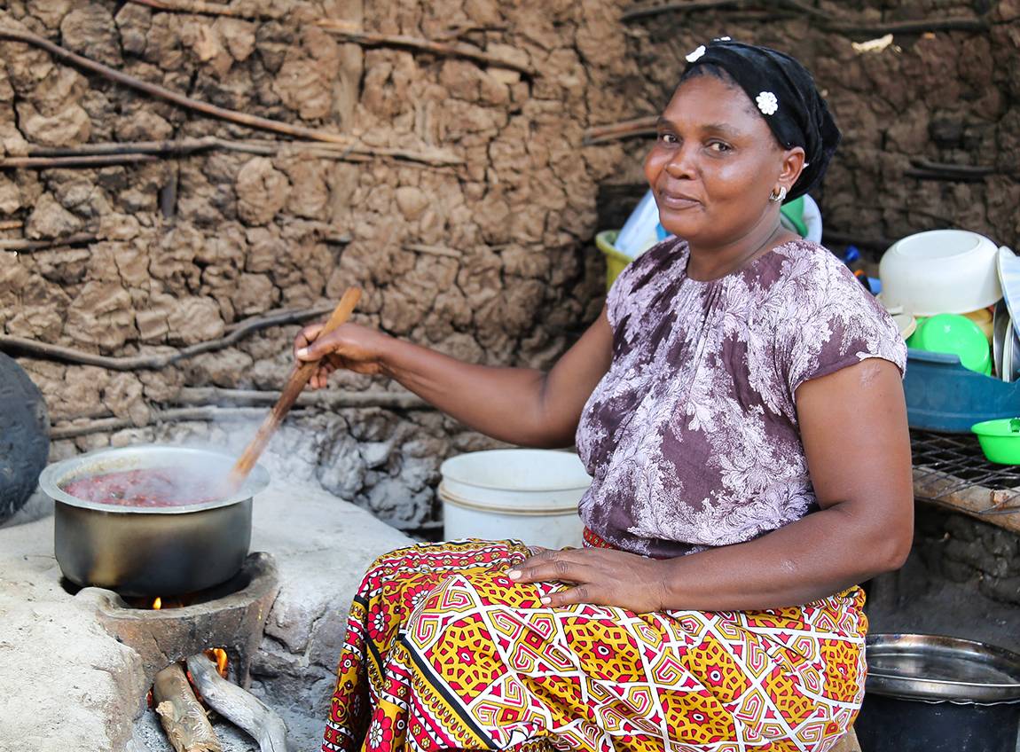 一名婦女坐在一些鍋子和碗盤中間，並用木勺子攪拌鍋裡的食物。